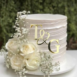 May include: A white wedding cake with gold glitter and a gold acrylic topper that says "T&G". The cake is decorated with white flowers.
