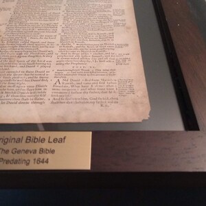 Puede incluir: Un primer plano de una p&aacute;gina enmarcada de una Biblia de Ginebra antigua, impresa antes de 1644. La p&aacute;gina est&aacute; amarillenta por el paso del tiempo y presenta texto en tinta negra. Una peque&ntilde;a placa dorada debajo de la p&aacute;gina dice "Hoja de Biblia original, La Biblia de Ginebra, Anterior a 1644."