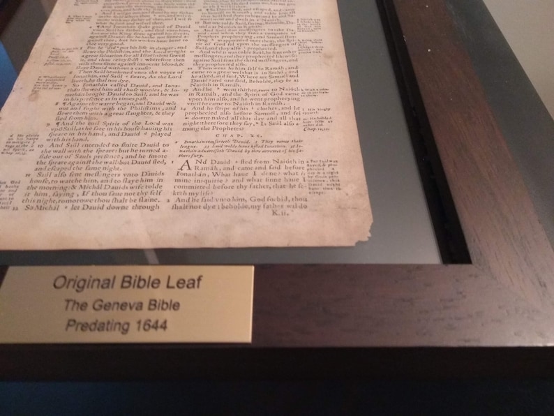 Puede incluir: Un primer plano de una p&aacute;gina enmarcada de una Biblia de Ginebra antigua, impresa antes de 1644. La p&aacute;gina est&aacute; amarillenta por el paso del tiempo y presenta texto en tinta negra. Una peque&ntilde;a placa dorada debajo de la p&aacute;gina dice "Hoja de Biblia original, La Biblia de Ginebra, Anterior a 1644."