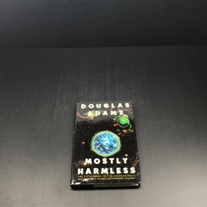 May include: A hardcover book titled "Mostly Harmless" by Douglas Adams. The cover features a dark background with a green alien, a blue and white swirling design, and the book's title in large, bold letters. The book is part of the Hitchhiker's Trilogy.