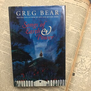 May include: A book cover with a blue and purple background featuring a fantasy landscape with a person riding a horse. The title of the book is "Songs of Earth and Power" by Greg Bear, the bestselling author of "Eon" and "Moving Mars".