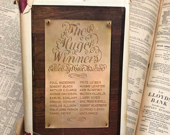 May include: A vintage book cover with a gold plaque that reads "The Hugo Winners Edited by Isaac Asimov". The plaque lists the names of Hugo Award winners, including Poul Anderson, Robert Bloch, Arthur C. Clarke, Avram Davidson, Samuel R. Delaney, Gordon Dickson, Harlan Ellison, Philip Jose Farmer, Daniel Keyes, Fritz Leiber, Murray Leinster, Anne McCaffrey, Walter M. Miller Jr., Larry Niven, Eric Frank Russell, Robert Silverberg, Clifford D. Simak, and Jack Vance.