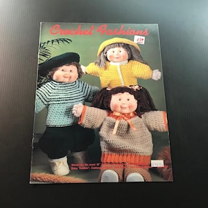 May include: A vintage magazine cover titled "Crochet Fashions" featuring three dolls dressed in crocheted outfits. The dolls wear a black beret, a yellow jacket, and a brown sweater. The magazine cover has a red title and includes text about wardrobe sizing.