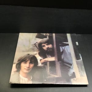 May include: Album cover featuring two men with long hair and beards. One man is leaning on a structure, while the other is in the foreground. The cover has a vintage aesthetic, with a slightly faded color palette.