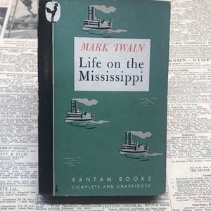 May include: A vintage paperback book titled "Life on the Mississippi" by Mark Twain. The cover is teal with illustrations of steamboats and the publisher's name, Bantam Books.