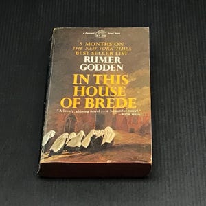 May include: A vintage paperback book titled "In This House of Brede" by Rumer Godden. The cover features text and an illustration of figures in white robes. The book is a former New York Times bestseller.