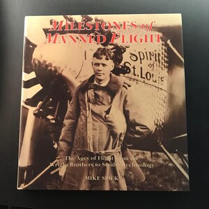 May include: A hardcover book titled Milestones of Manned Flight by Mike Spick. The sepia-toned cover features a photograph of a pilot standing in front of the Spirit of St. Louis airplane, with the subtitle The Ages of Flight from the Wright Brothers to Stealth Technology.