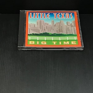 May include: A CD case featuring the album "Little Texas Big Time." The cover art displays a stylized cityscape in shades of gray, green, and orange, with the band's name in red and blue. The CD case is black and clear.