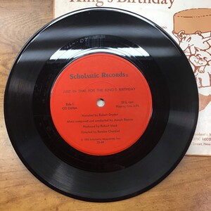 Peut inclure: Un disque vinyle noir avec une &eacute;tiquette rouge. L'&eacute;tiquette indique "Scholastic Records" et "Just in Time for the King's Birthday". Le disque tourne &agrave; 33 1/3 tours par minute et a une dur&eacute;e de lecture de 6:55.