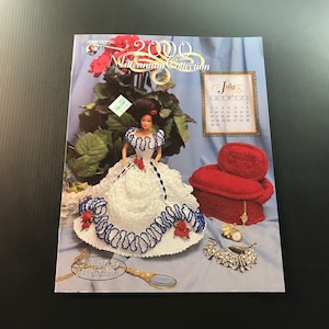 May include: A magazine cover featuring a doll in a white dress with blue and red accents, a red chair, and a July calendar. The text reads "2000 Millennium Collection" and "Anne's Attic Original."