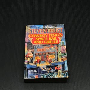 May include: A paperback book titled "Cowboy Feng's Space Bar and Grille" by Steven Brust. The cover features a colorful illustration of a futuristic bar with the word "BAR" above the entrance. The text "It's the best bar on Earth... and everywhere else" is also visible.