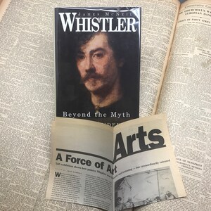 May include: A book titled "Whistler: Beyond the Myth" by James McNeil, featuring a portrait of the artist on the cover. The book is open to a page with the text "A Force of Art" and a smaller image of a painting.