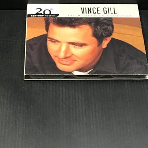 May include: A CD case featuring Vince Gill, with the text "20th Century Masters" and "The Millennium Collection." The cover displays a close-up of Vince Gill's face against a warm, neutral background. The CD case is on a dark surface.