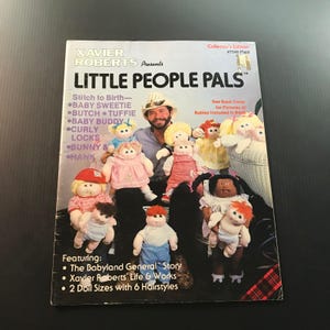 May include: A vintage magazine cover for "Little People Pals" by Xavier Roberts. The cover features a man surrounded by various dolls, with text including "Baby Sweetie" and "Baby Buddy." The magazine is a collector's edition.