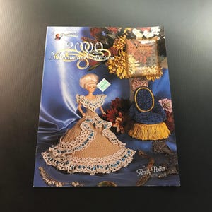 May include: A magazine cover featuring a doll in a crocheted gown, with the text "2000 Millennium Collection." The doll has blonde hair and is wearing a tan dress with blue accents. A small blue and gold decorative piece is also visible.