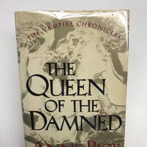 May include: A book cover with a gold background and a black and white image of a woman's face. The title of the book is "The Queen of the Damned" by Anne Rice. The book is part of "The Vampire Chronicles" series.