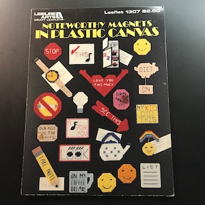 May include: A black leaflet titled "NOTEWORTHY MAGNETS IN PLASTIC CANVAS" featuring various cross-stitch designs. Designs include a stop sign, watch, smiley faces, and phrases like "LOVE YOU THIS MUCH" and "TAKE NOTE".