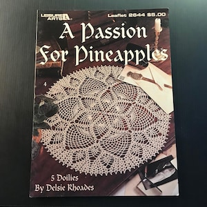 May include: A book cover titled "A Passion for Pineapples" featuring a cream-colored doily design. The cover also includes the text "5 Doilies By Delsie Rhoades" and the Leisure Arts logo.