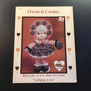 May include: A vintage craft pattern titled "Oatmeal Cookie" featuring a doll with a crocheted hat, dress, and mittens. The pattern is from "Lollipop Lane" and includes a price tag. The doll has a smiling face and is wearing black shoes.