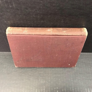 May include: A vintage book with a burgundy cover and a tan spine. The spine has faded gold lettering. The book's edges show signs of wear, suggesting its age. The book is positioned on a dark surface.