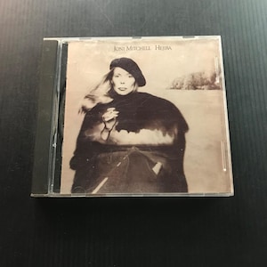 May include: CD case featuring the album "Hejira" by Joni Mitchell. The album cover is a sepia-toned photograph of a woman wearing a beret and dark clothing. The album title and artist's name are printed at the top.