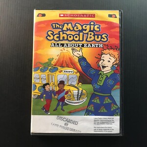 May include: A VHS tape of "The Magic School Bus: All About Earth" featuring a yellow school bus with a volcano erupting in the background. The cover has the Scholastic logo and a cartoon of a woman in a blue dress.