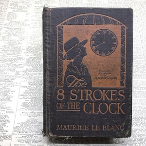 May include: A vintage hardcover book titled "The 8 Strokes of the Clock" by Maurice Le Blanc. The cover features a silhouette of a woman wearing a hat, looking at a clock face with Roman numerals. The text "The Latest Exploits of Arsene Lupin" is printed in gold lettering.