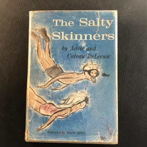May include: A hardcover book titled The Salty Skinners by Adèle and Cateau DeLeeuw, illustrated by Arnold Spilka. The blue cover features a sketch of two people swimming underwater wearing masks and fins, with a small fish visible near the top figure.
