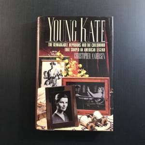 May include: A hardcover book titled "YOUNG KATE" with the subtitle "THE REMARKABLE HEPBURNS AND THE CHILDHOOD THAT SHAPED AN AMERICAN LEGEND" by Christopher Andersen. The cover features framed photos and a floral arrangement. The book is a biography.