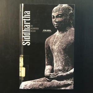 May include: A book titled "Siddhartha" by Hermann Hesse. The cover features a grayscale image of a seated Buddha statue. The book is published by Perma-Bound. The title is in white, vertical text on the left side.