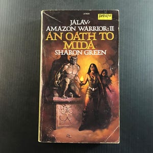 May include: A mass market paperback book titled Jalav: Amazon Warrior: II An Oath to Mida by Sharon Green. The cover features a dark fantasy illustration of a horned creature sitting on a stone pedestal next to a woman holding a torch.