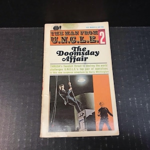May include: Vintage paperback book titled "The Man From U.N.C.L.E. 2: The Doomsday Affair." The cover features a man in a suit swinging from a chain, with orange and white text and illustrations. The book is slightly worn.