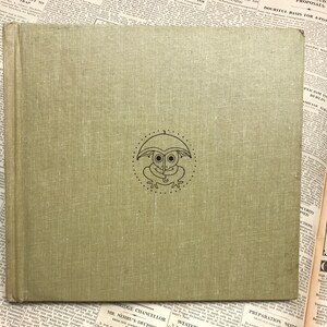 May include: A green hardcover book with a frog wearing an umbrella on the front cover. The frog is outlined in black and has a white face.