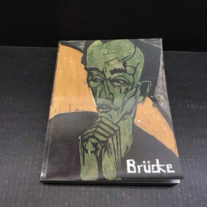 May include: A book cover featuring a green-toned portrait of a man with his hand on his chin. The background has a gold and black color scheme, with the word "Brücke" in white text.