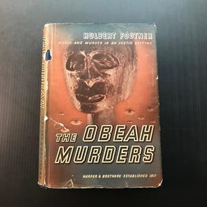 May include: Vintage book cover for "The Obeah Murders" by Hulbert Footner. The cover features a tribal mask and multiple eyes against an orange and blue background. The title is in bold white letters.