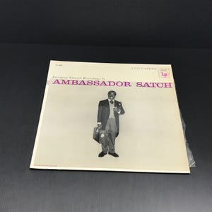 May include: A vintage LP record album cover featuring a black and white image of a man in a suit holding a briefcase and a drink. The album is titled "Ambassador Satch" with the Columbia Records logo.