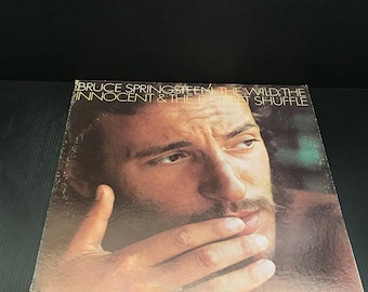 May include: Vintage vinyl record album cover featuring Bruce Springsteen. The album cover has a sepia-toned portrait of Springsteen with the album title "The Wild, The Innocent & The E Street Shuffle" printed at the top.
