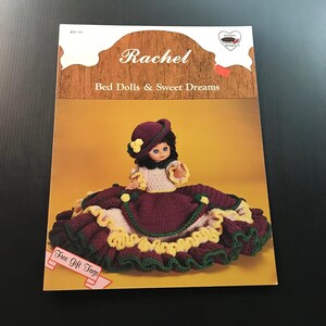 May include: A vintage craft pattern book titled "Rachel Bed Dolls & Sweet Dreams." The cover features a crochet doll in a burgundy dress with yellow and green accents. The doll has a matching hat and a white top.