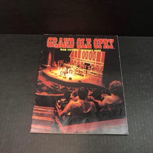 May include: A vintage "Grand Ole Opry" history book. The cover features the title in red, with a stage scene and audience. The book is a WSM Picture History Book.
