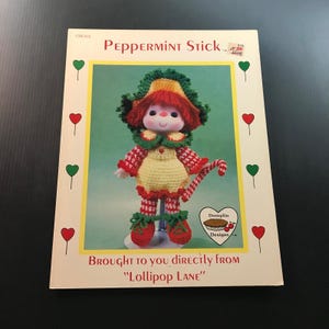 May include: A vintage crochet pattern book titled "Peppermint Stick." The cover features a doll in a crocheted outfit with a candy cane. The doll has red hair and is wearing a yellow dress with red and white accents. The text reads "Brought to you directly from 'Lollipop Lane'"