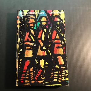 May include: A hardcover book titled "In Flanders Fields" by Leon Wolff. The cover features a colorful, abstract illustration of three figures in a stained-glass style, with text in white and yellow. The book is set against a dark background.