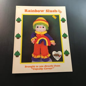 May include: A vintage pattern cover titled "Rainbow Slush" featuring a crocheted doll in orange pants, a rainbow-shaped vest, and a yellow and red hat. The text "Brought to you directly from 'Cupcake Corner'" is also visible.