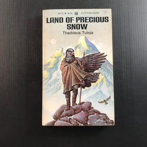 May include: A paperback book titled Land of Precious Snow by Thaddeus Tuleja. The cover features an illustration of a bird-headed figure in a cloak standing on a rocky outcrop before snow-capped mountains, with a small bird flying nearby.