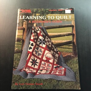 May include: A beginner's guide to quilting book titled "Learning to Quilt." The cover features a colorful quilt with red, white, and blue geometric patterns draped over a wooden fence. The book's title is in large, bold letters.