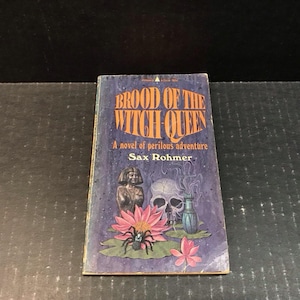 May include: Vintage paperback book titled "Brood of the Witch Queen" by Sax Rohmer. The cover features a skull, a spider, a lotus flower, and an illustration of a statue. The background is a dark blue with a purple hue.