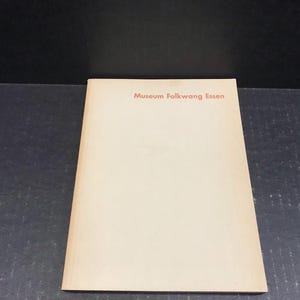 May include: A beige booklet with the text "Museum Folkwang Essen" in red lettering. The booklet is slightly angled, resting on a dark surface. The background is black.