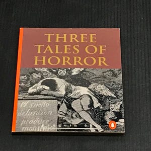 May include: A paperback book titled "THREE TALES OF HORROR" with a red and orange spine. The cover features a black and white illustration of a person lying down with owls and cats. The text "El sueno de la razon produce monstruos" is also visible.