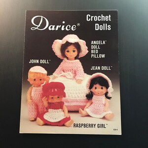May include: A vintage Darice crochet dolls pattern book. The cover features four crochet dolls: Angela, John, Jean, and Raspberry Girl. The dolls are wearing crochet outfits in pink and red. The book is on a black background.