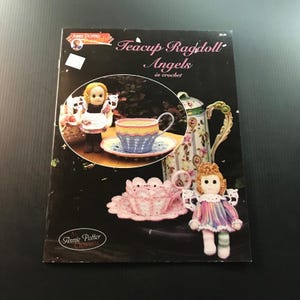 May include: A black book cover with the title "Teacup Ragdoll Angels in crochet." The cover features crochet angel dolls, a teacup, and a saucer. The teacup is blue, yellow, and pink. The dolls have wings and colorful dresses.