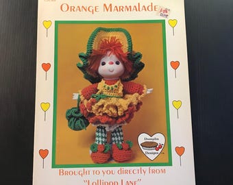 May include: A vintage book cover featuring a crochet doll named "Orange Marmalade." The doll has a red and yellow dress, green hat, and a green bag. The text reads "Brought to you directly from Lollipop Lane."